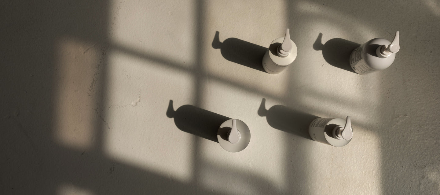 Four Basic Maintenance product bottles arranged on the floor between the shadows cast by a window.